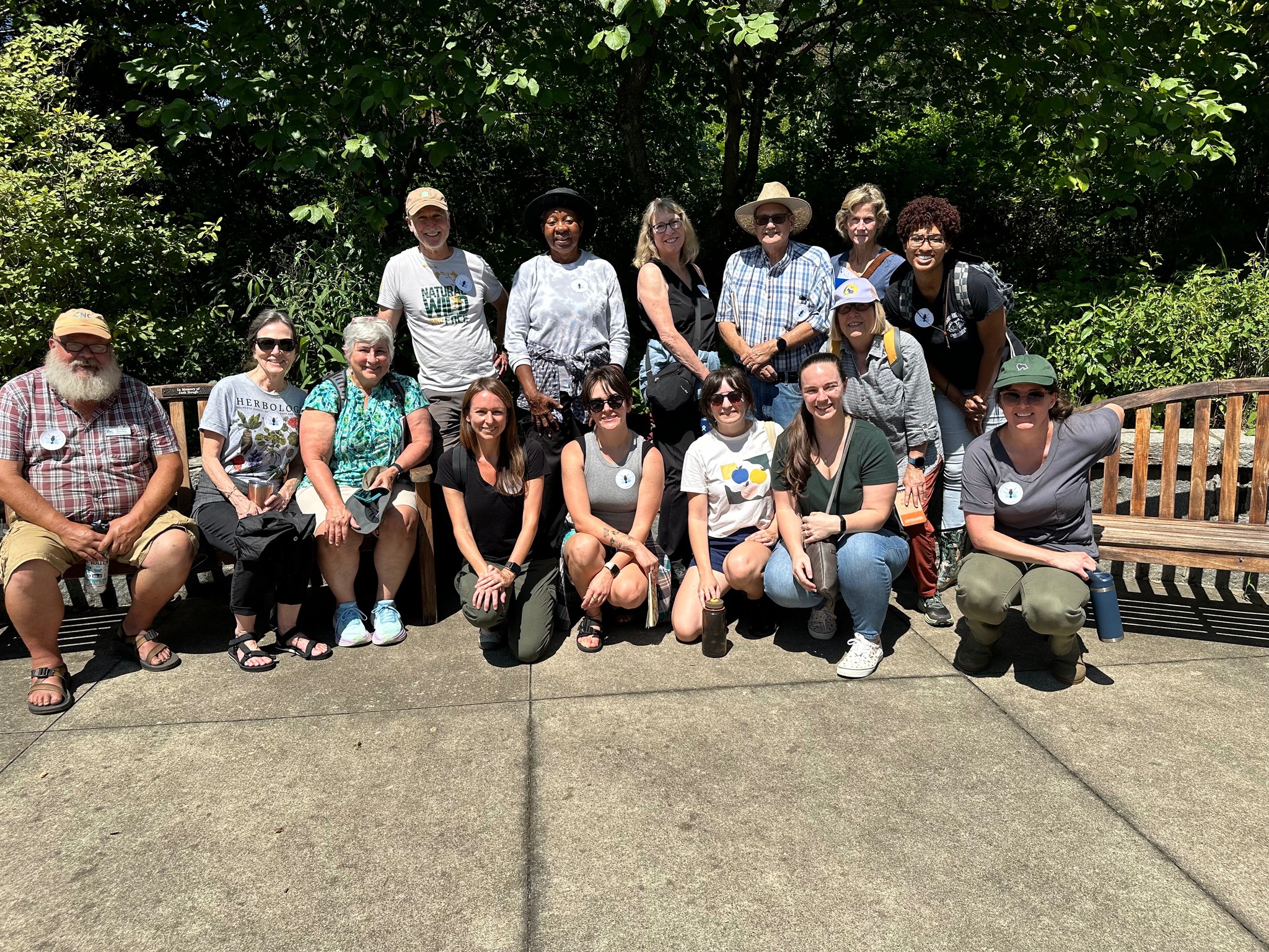 15 smiling people stand and sit in a group outside on a sunny day.