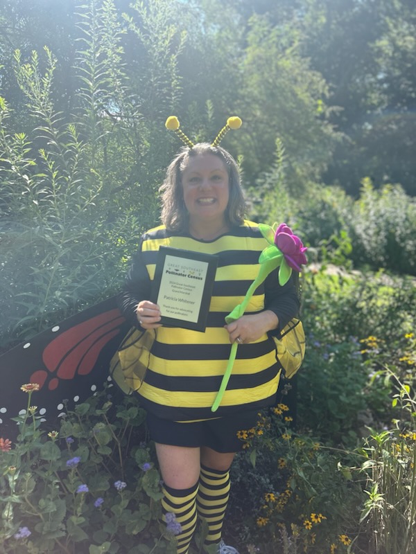 A person in a bee costume holding a placard and an oversized flower, standing outdoors.