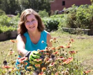 Photo of a person in a blue shirt in a garden, smiling while reaching for flowers.