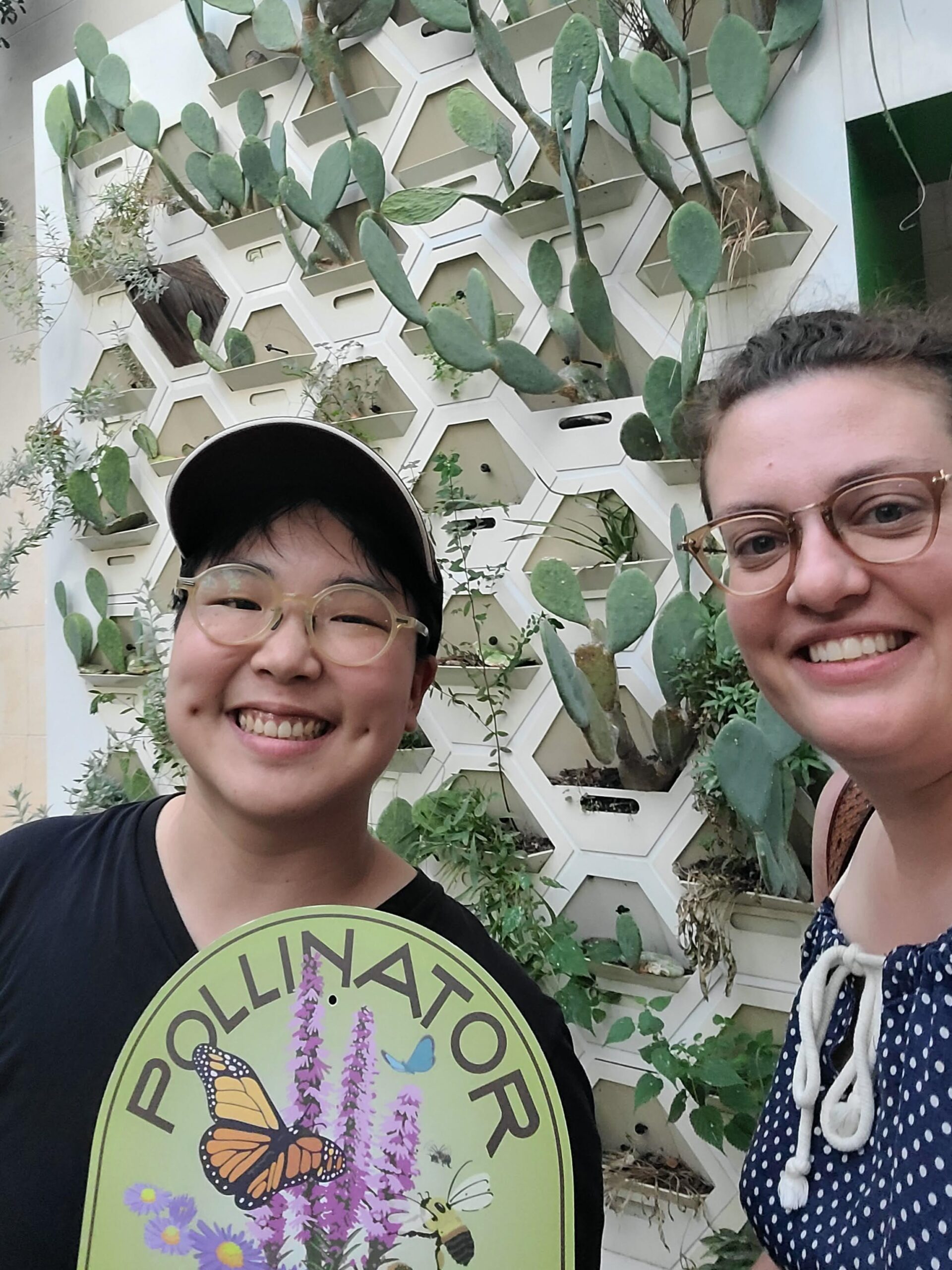 Two people smile holding a pollinator sign with plants on a wall in the background.