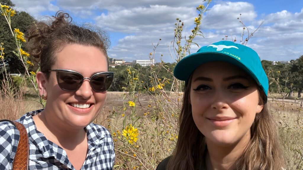 Two people smile on a sunny day with yellow flowers and yellow grass in the background.