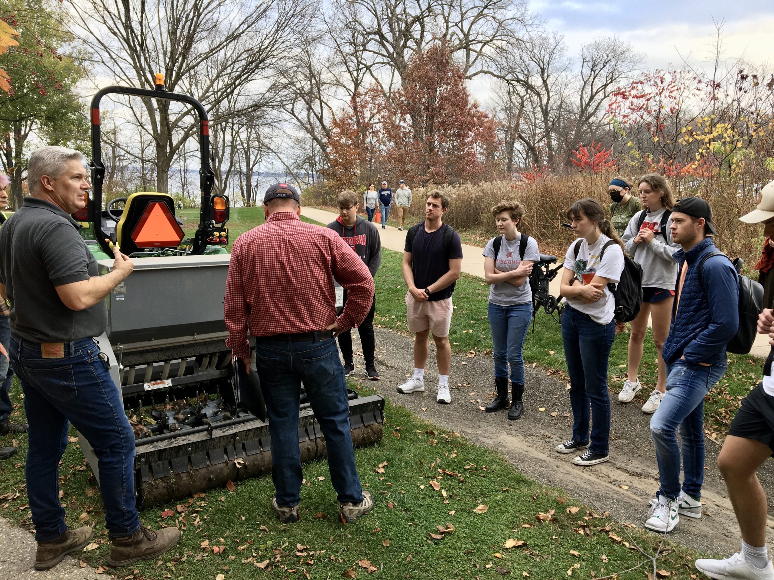 Students stand around tractor as a grounds staffer in a red shirt instructs them