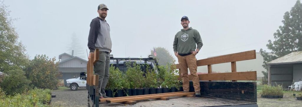 Two men standing in the bed of a pickup truck filled with green plants