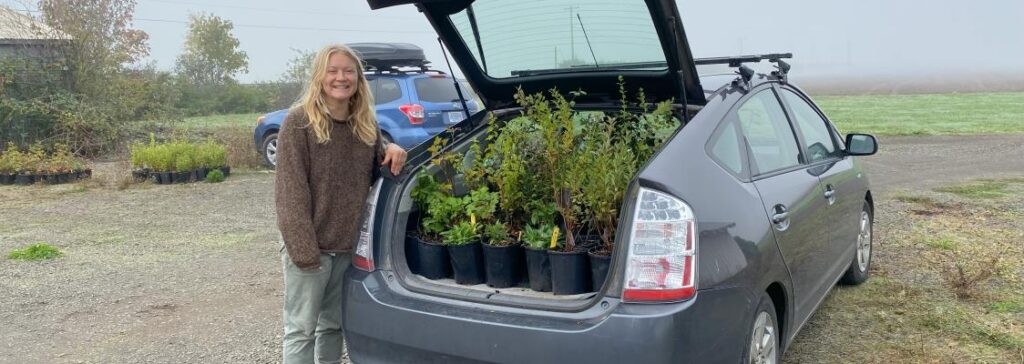 A woman stands smiling beside her car with an open trunk full of green plants