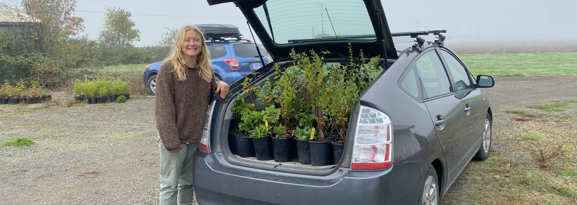 A woman stands smiling beside her car with an open trunk full of green plants