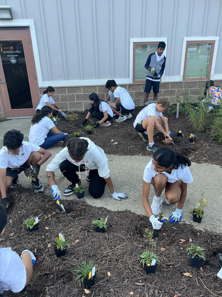 10 kids crouch down in a garden, planting small green plants in front of a building.