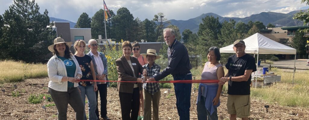 Nine people in a row hold a red ribbon in front of a garden.