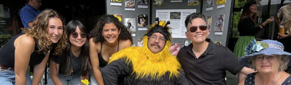 6 people, 1 in a fuzzy bee costume, pose in front of a poster board.