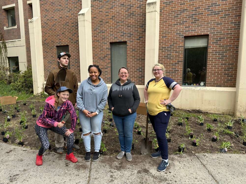 Four people pose smiling in front of plants laid out for planting. in front of a red brick building. 