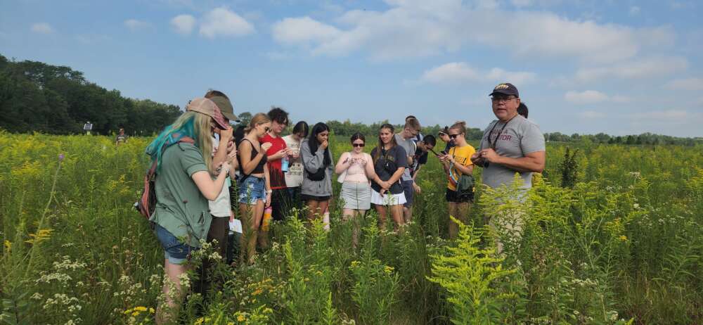 Around a dozen people stand in a flowering prairie.