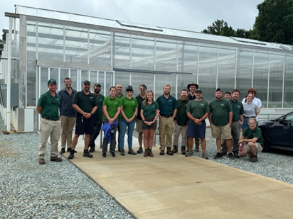 A line of a bout 18 people smile in front of a large greenhouse.