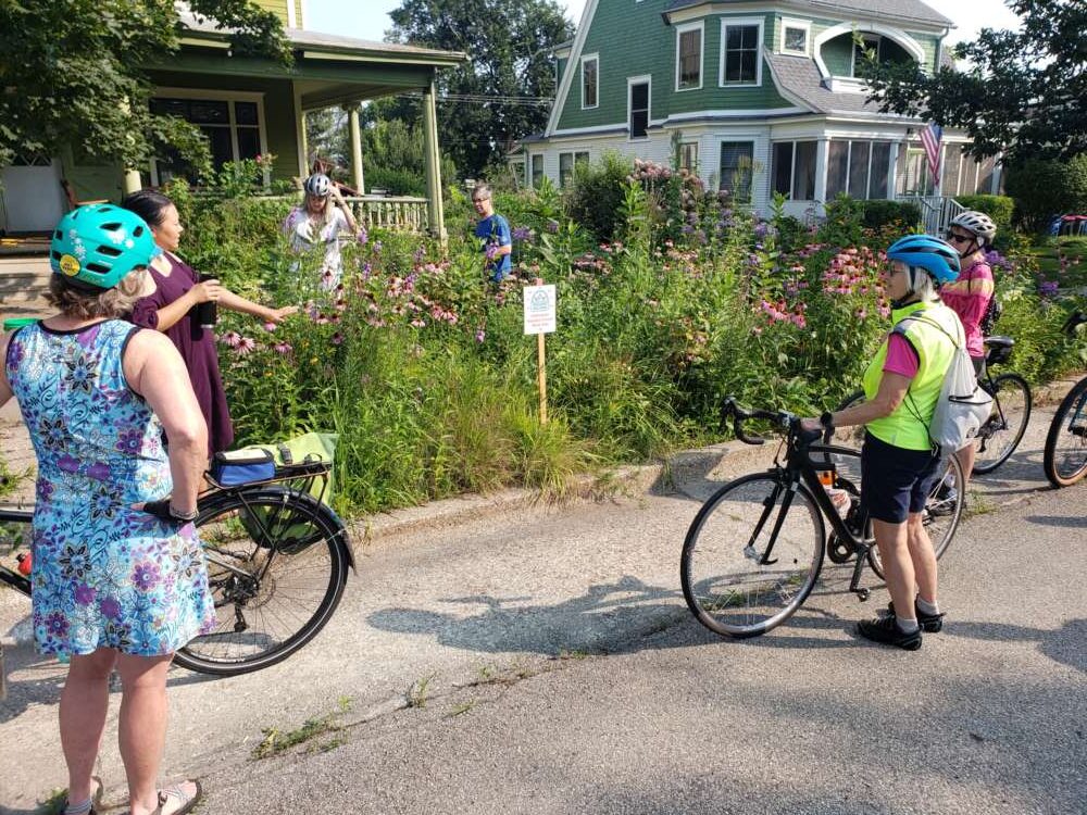 6 people, some on bikes, look towards a yard with flowering plants.