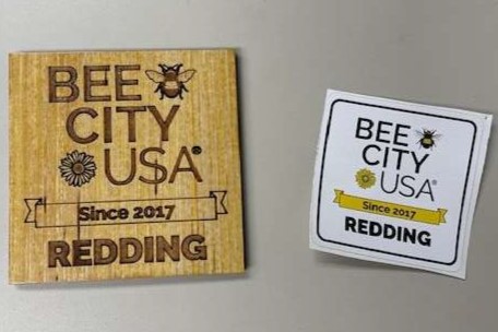 A sticker and a wood badge reading Bee City USA Redding.