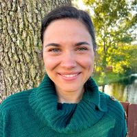 A person in a teal sweater smiles with a tree in the background.