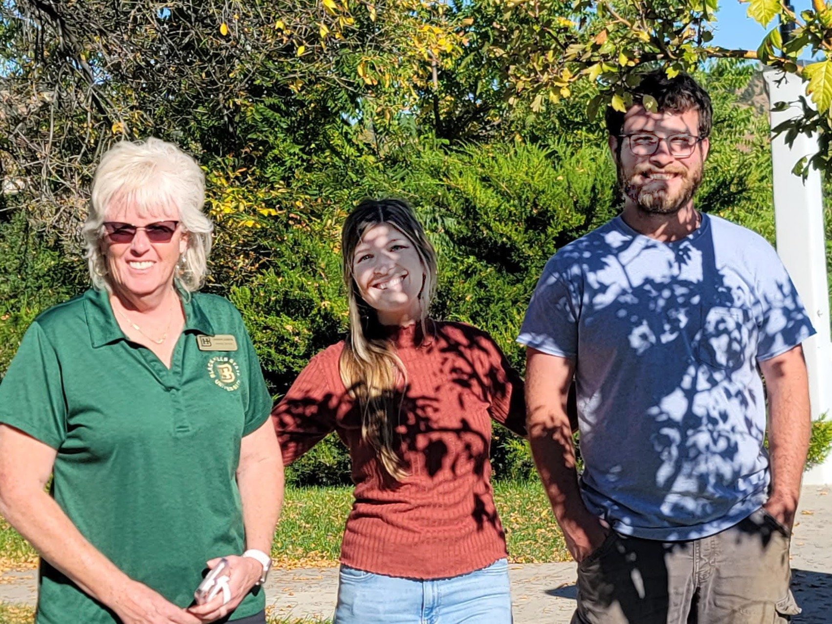 Three people smile in the shadow of a tree.