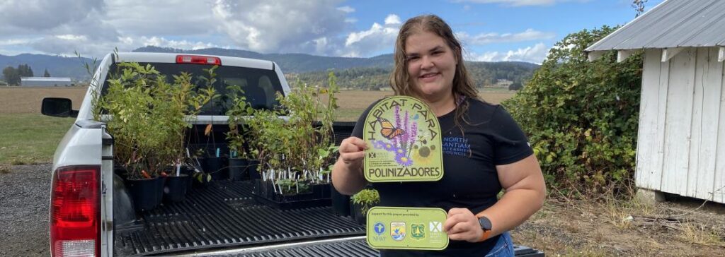 A person jolds up a green pollinator habitat sign in front of a full truck bed of plants