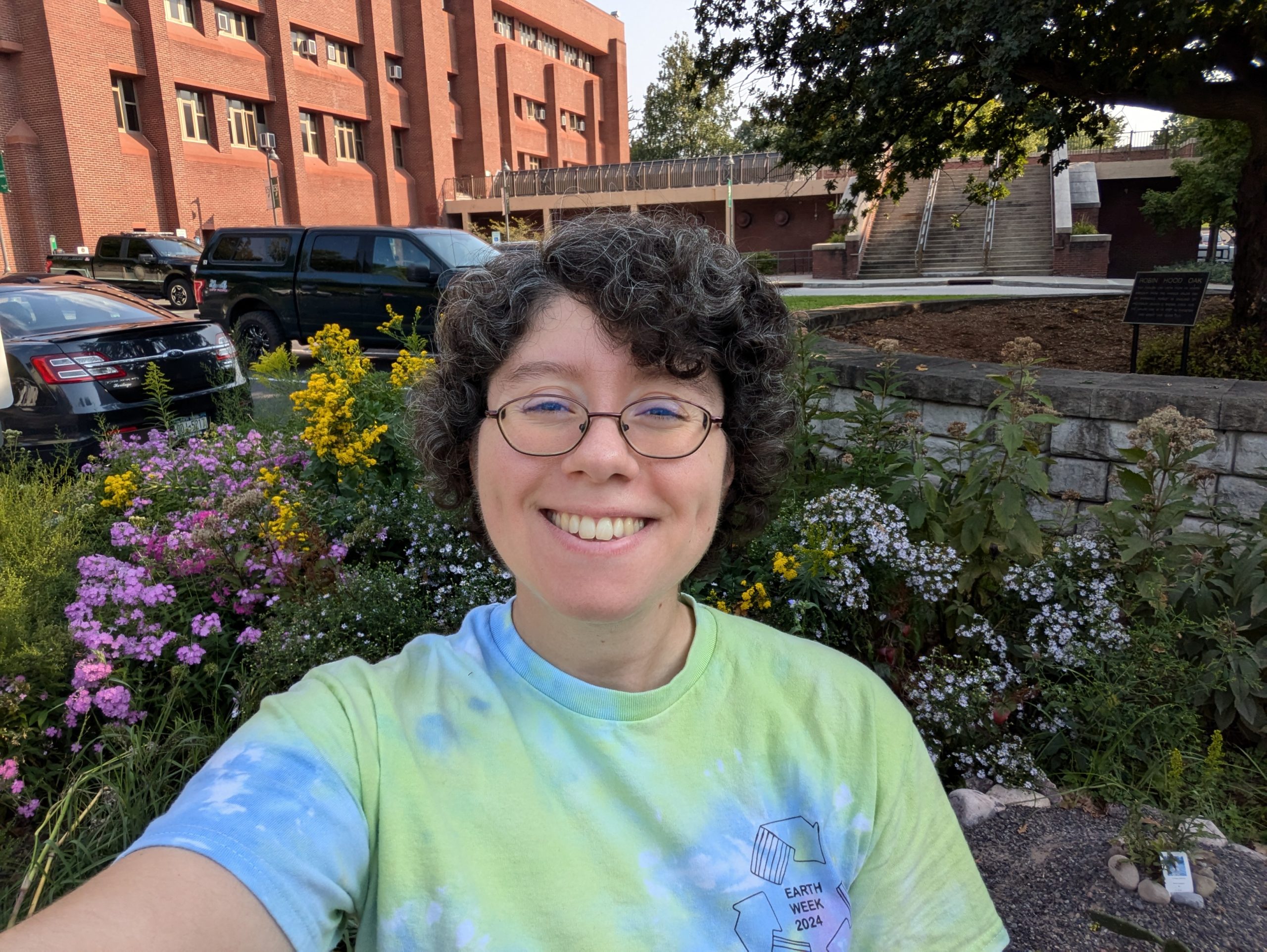 A person in a green and blue short smiles into the camera, in front of colorful blooming flowers.