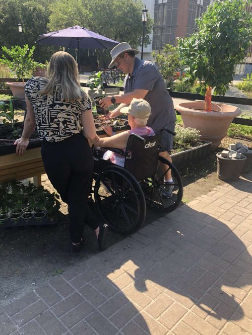 Two people stand and one person sits in a wheelchair, facing a raised garden bed with plants.