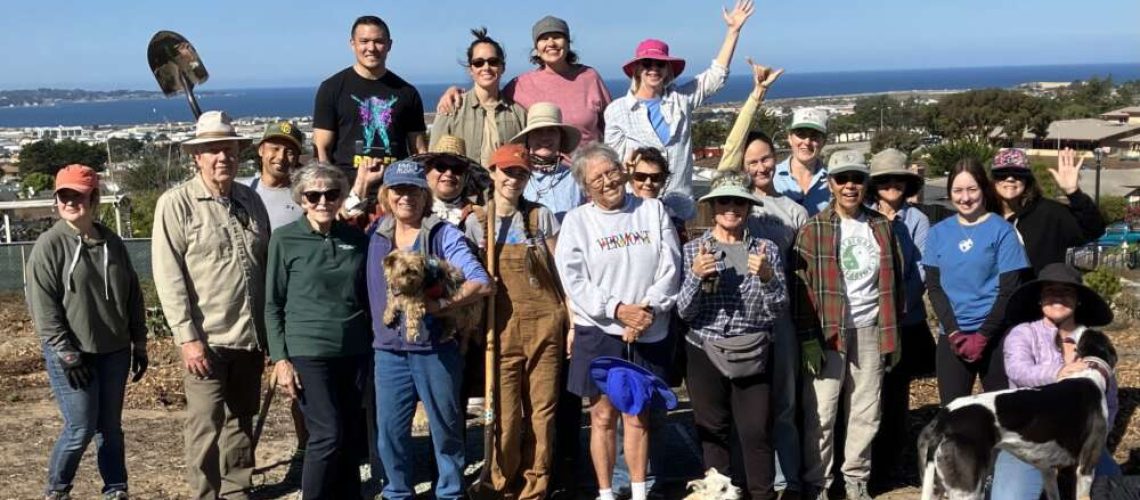 A large group of people pose close together on a sunny day with the ocean in the background.