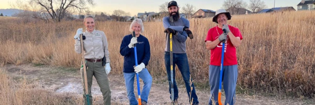 Four people stand in a row holding shovels in a field with tall golden grass with houses in the background.