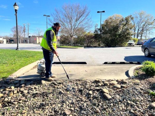 A person in a neon green vest holds a long metal pole, flame-weeding on gravel.