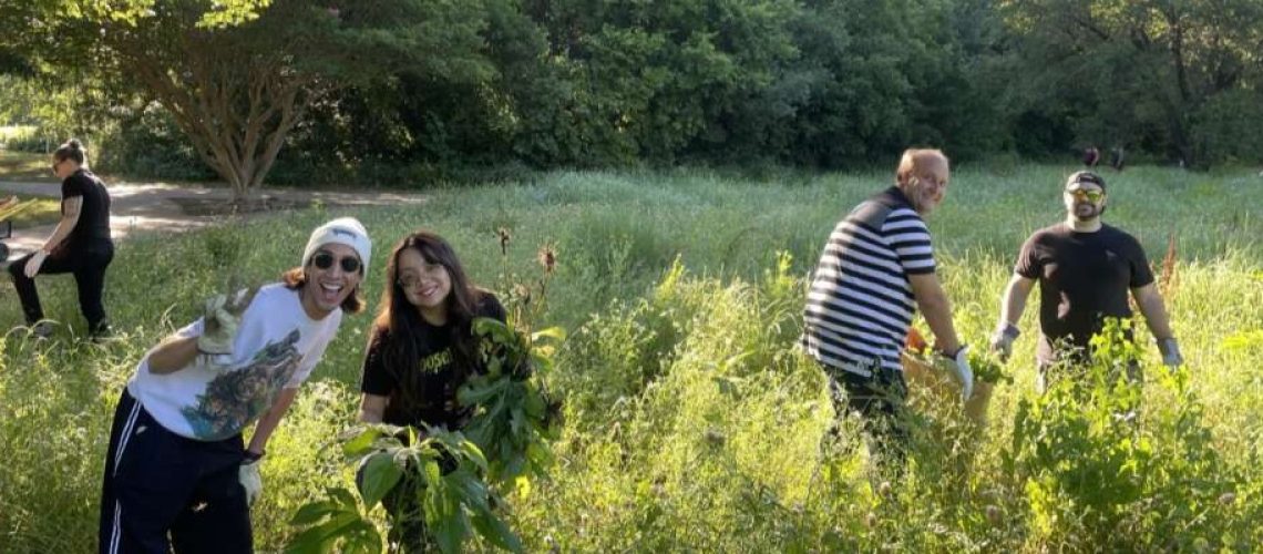 Groups of smiling people work in a sunny green meadow at the camera. A dark green forest is in the background.