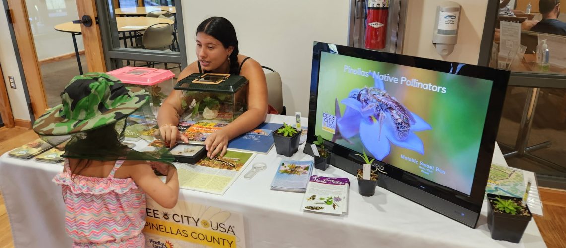 An adult sits behind a display table talking to a child in a beekeeping hat.
