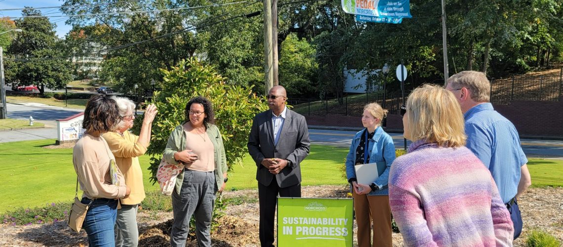 A circle of people listen to a speaker on a sunny day. A green yard sign in the middle of the group reads "Sustainability in Progress".