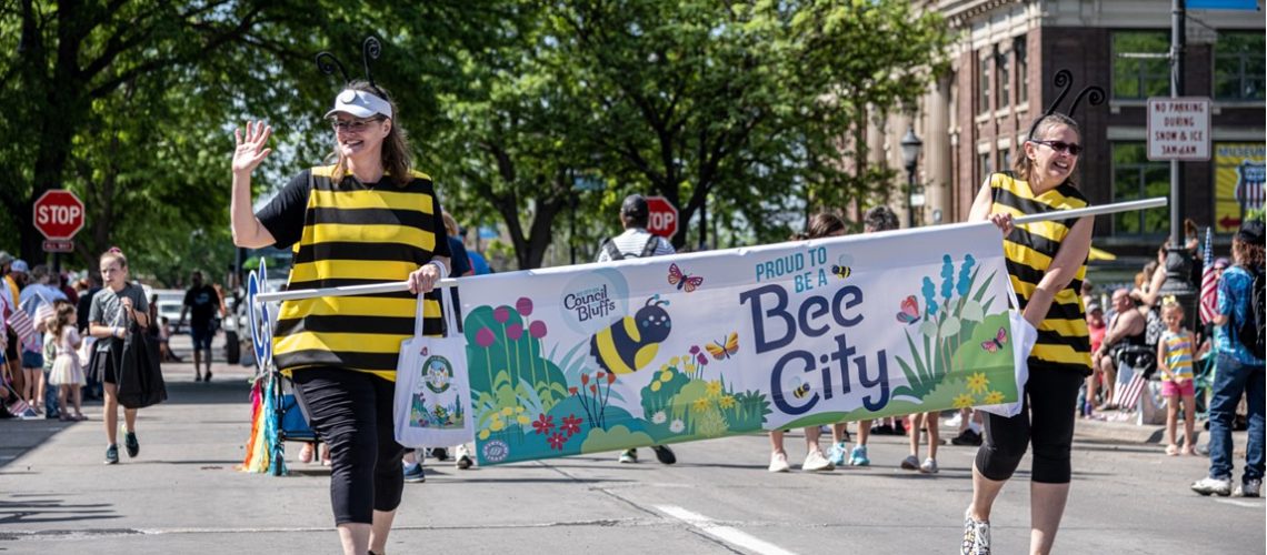 Two people in bee costumes hold a large Bee City banner walking down a street in a parade.