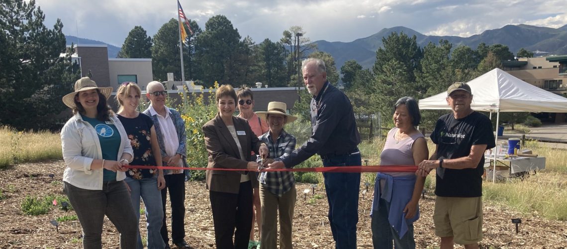 Nine people in a row hold a red ribbon in front of a garden.