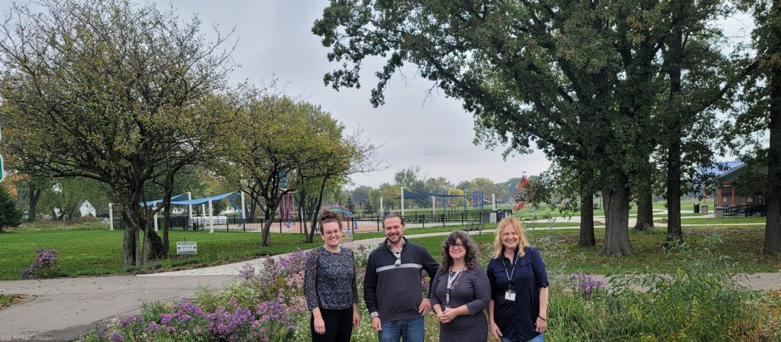 Four adults in darker clothes stand in a row in from of flowers with trees and a playground in the background.