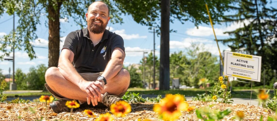 A smiling person in a dark polo shirt sits cross-legged outside in front of red and yellow blanket flowers with a sign reading "Active Planting Site".