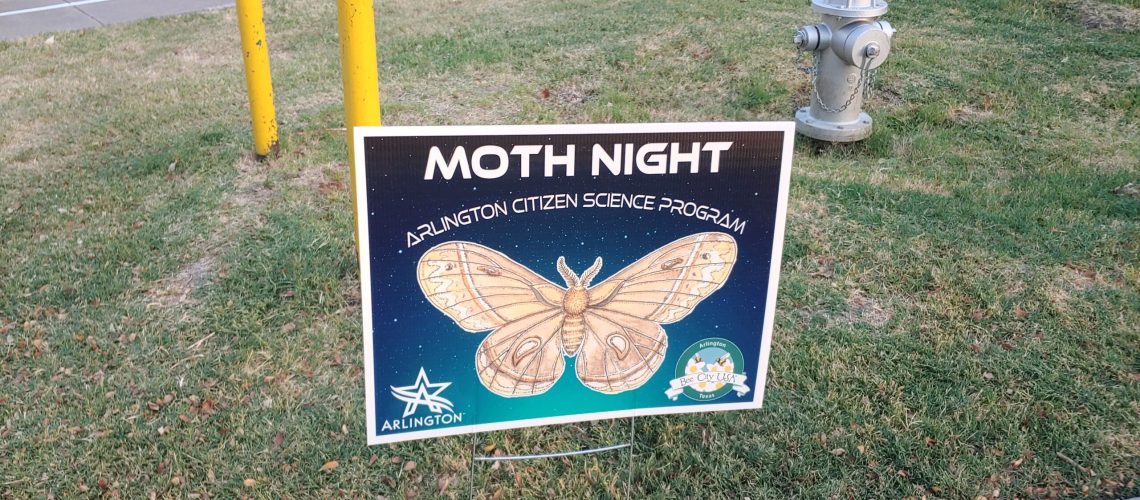 A dark blue lawn sign saying Moth Night with a picture of a moth, on a green lawn