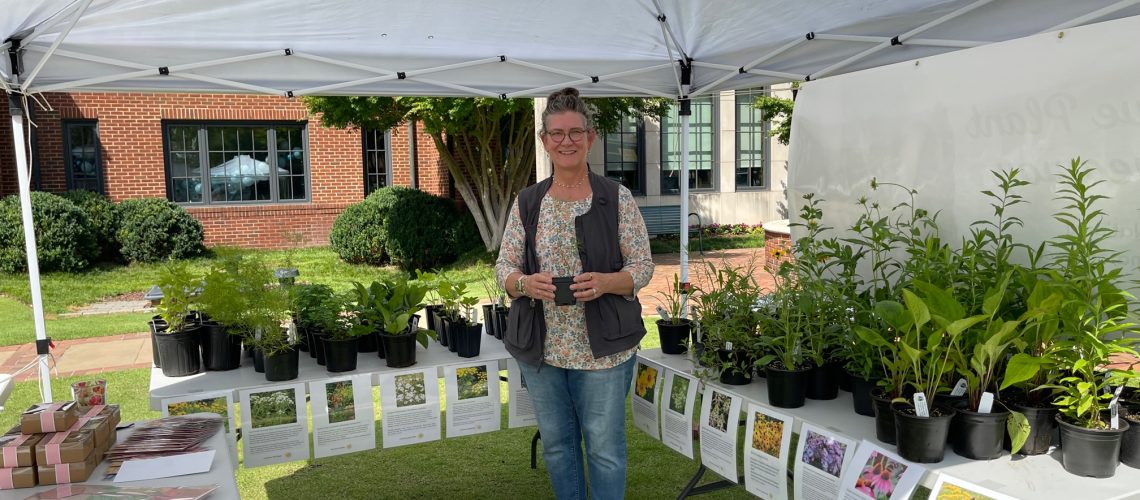 A person in a dark vest and floral shirt stands holding a small plant pot. Standing under a white outdoor canopy, they are surrounded by rows of green plants in black pots on white tables. There is a red brick building in the background.