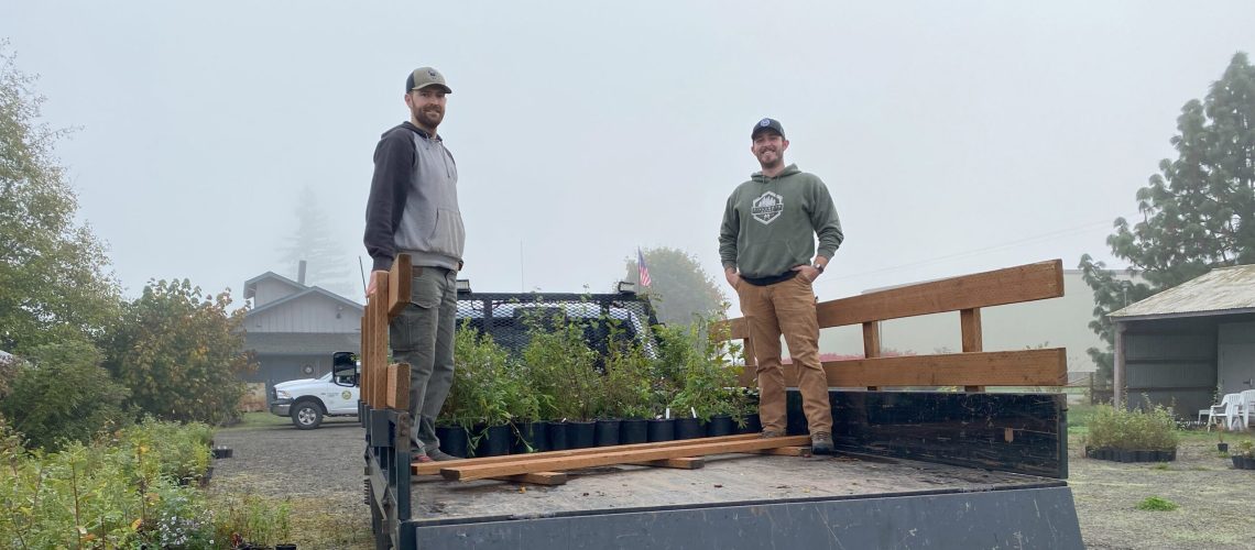 Two smiling people in hoodies stand in the bed of a pickup truck, with a flat of green plants in black pots behind them.