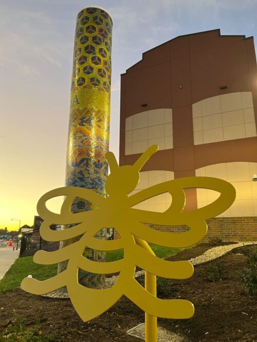 A shiny mosaic column at sunset, with a yellow metal bee cutout in the foreground and a red brick building in the background.
