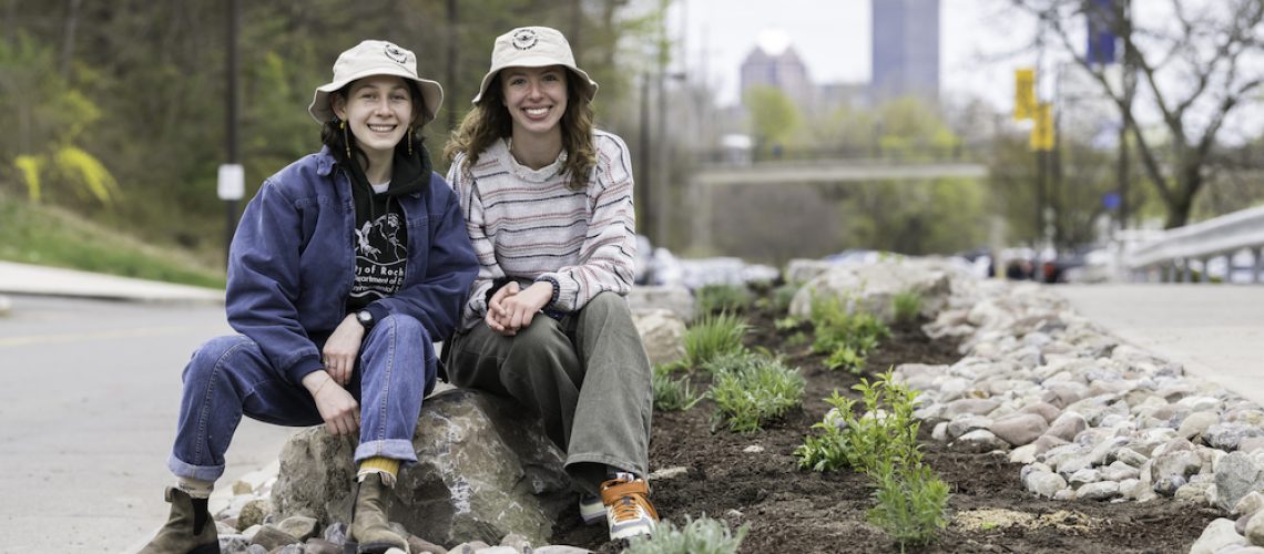 Two university students in tan bucket hats smile, sitting on a rock next to a strip of plants and rocks along a road with trees and tall buildings in the background.